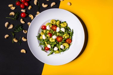 Mozarella, marul, roka, domates ve pesto soslu makarna sarı-siyah bir tabakta.