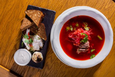 Borscht with meat, potatoes, parsley, beets, and cabbage in a plate on the table with slices of bread, bacon, pea sprouts, sour cream in a saucepan and fried garlic on a stone plate