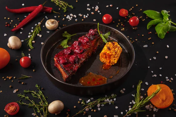 Fried pork steak with sauce, cherries, parsley and corn on an iron plate with mushrooms, peas, red chili peppers, salt, tomatoes and basil on a black background