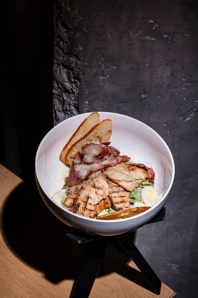 Grilled fried meat with bacon, boiled egg, parmesan, bread and sauce in a plate on a stand on the edge of the table against the background of a stone wall