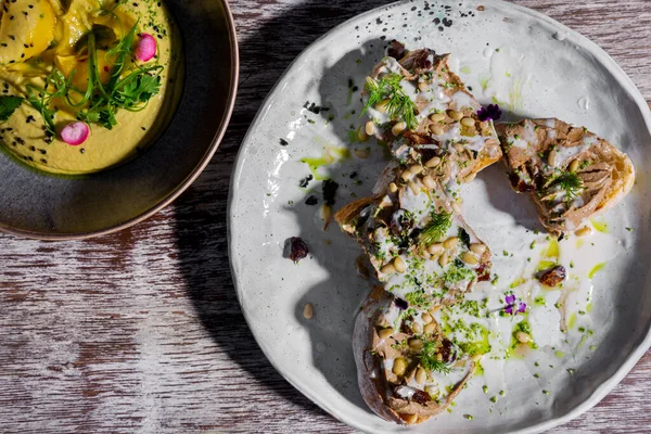 Sandwich with meat, wheat, flower petals, dill, and sauce in a plate on a wooden table and hummus in another plate