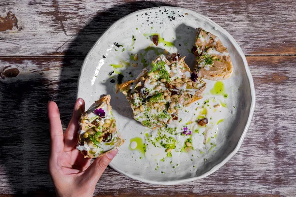 Sandwich with meat, wheat, flower petals, dill, and sauce in a plate on a wooden table with human hands