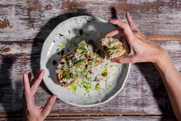 Sandwich with meat, wheat, flower petals, dill, and sauce in a plate on a wooden table with human hands