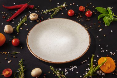 White plate stands on a dark background with mushrooms, peppercorns, red chili peppers, salt, tomatoes and basil on a black background