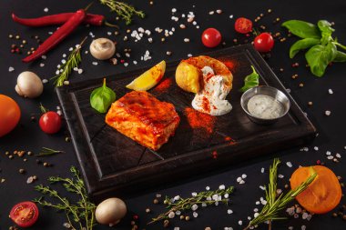 Fried salmon steak with fried potatoes, sauce, pepper, lemon and basil on an iron plate with mushrooms, peas, red chili peppers, salt, tomatoes and basil on a black background