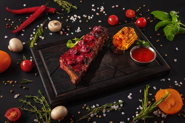 Fried pork steak with sauce, cherries, parsley and corn on a wooden board with mushrooms, peas, red chili peppers, salt, tomatoes and basil on a black background