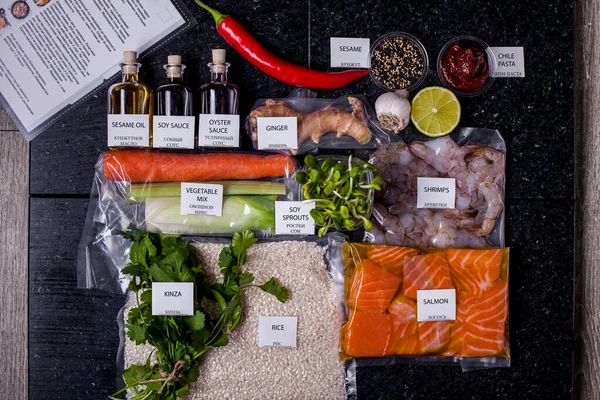Cooking ingredients. Sesame oil, soy and oyster sauce in a glass jar, salmon, rice, vegetable mix, soy sprouts, ginger, shrimps in a plastic bag, sesame and chili paste in a plastic glass are on a wooden table with a recipe book, lemon, garlic and ci