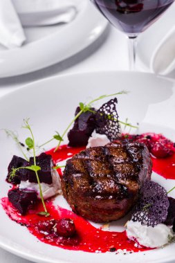 Beyaz yuvarlak bir tabakta, lahanalı, pancarlı, dantelli, siyah cipsli ve vişne soslu ızgara biftek ve bir bardak kırmızı şarap. Dikey yönelim 