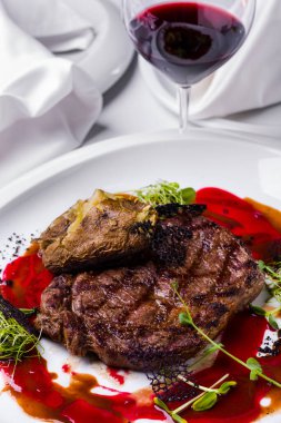 Bir parça ızgara biftek, lahana salatası, fırında patates, pancar, dantelli siyah cips ve bir bardak kırmızı şarapla birlikte beyaz yuvarlak bir tabakta vişne sosu. Dikey yönelim