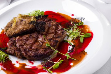 Bir parça ızgara biftek, lahana salatası, fırında patates, pancar, dantelli siyah cips ve beyaz yuvarlak tabakta vişne sosu. Yatay yönelim