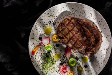 Kemiğin üzerinde bir parça ızgara biftek lahanası, kumkuat, turp, salatalık, mung bifteği salatası, siyah kumaş arka planda beyaz yuvarlak bir tabakta kırmızı biber. Yatay yönelim 