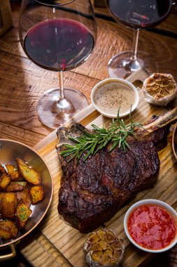 Grilled beef steak on the bone with spices, garlic, pepper, rosemary, red and white sauce, fried potatoes on a wooden board with cutlery, glasses of red wine, pepper shaker and salt shaker. Vertical orientation 