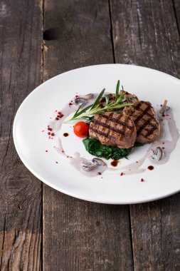 Grilled pork steak, rosemary, cherry tomato, red peppercorns, mushroom sauce, spinach on a round plate and a wooden table. Vertical orientation 
