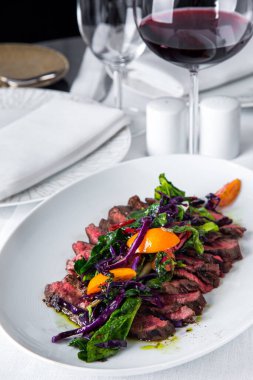 Grilled pork steaks with purple salad, kumquat, red chopped chili and spinach on a white oval plate with a glass of red wine. Vertical orientation 