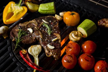 Grilled beef steak on the bone with rosemary, garlic, onion, pepper, zucchini, tomatoes lying on the embers. Horizontal orientation 
