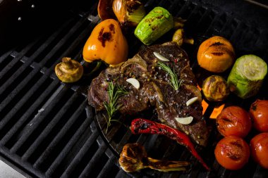 Grilled beef steak on the bone with rosemary, garlic, onion, pepper, zucchini, tomatoes lying on the embers. Horizontal orientation 