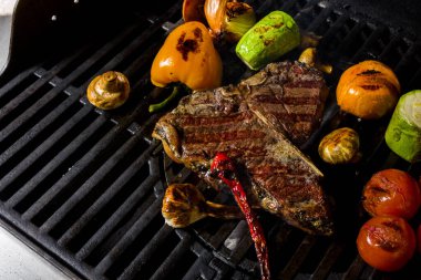 Grilled beef steak on the bone with rosemary, garlic, onion, pepper, zucchini, tomatoes lying on the embers. Horizontal orientation 