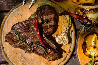 Grilled beef on the bone with rosemary, garlic, onion, pepper, zucchini, tomatoes lying on a round wooden board with salt, pepper and a meat fork Horizontal orientation 