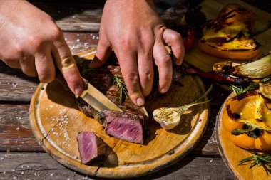 Grilled beef on the bone with rosemary, garlic, onion, pepper, zucchini, tomatoes lying on a round wooden board with salt, pepper, cut with a knife while holding hands. Horizontal orientation 