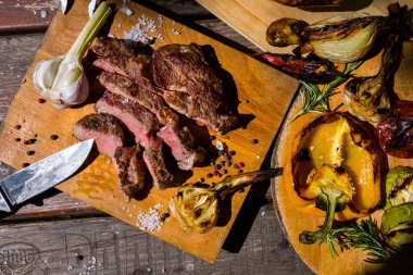 Roast beef on the bone with rosemary, garlic, onions, peppers, zucchini, tomatoes on a round wooden board, with salt, pepper, and a meat knife. Horizontal orientation