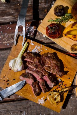 Roast beef on the bone with rosemary, garlic, onions, peppers, zucchini, tomatoes lying on a round wooden board, with salt, pepper, a fork and a meat knife. Vertical orientation 