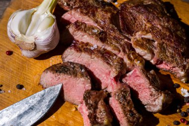 Roast beef on the bone with rosemary, garlic, onions, peppers, zucchini, tomatoes on a round wooden board, with salt, pepper, and a meat knife. Horizontal orientation