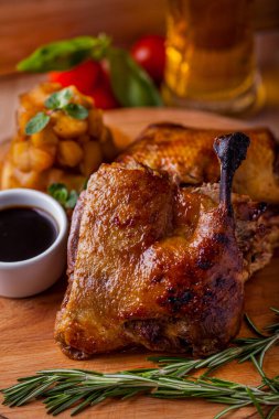 Grilled duck leg on the bone with rosemary, potatoes, basil and tomatoes with sauce on a wooden board with a glass of beer. Vertical orientation