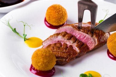 Roast duck fillet with fork, knife, sauce, balls and sprouts of green peas on a round plate. Horizontal orientation