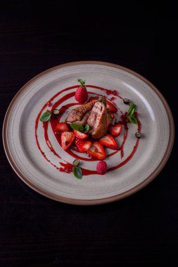 Grilled duck pieces in sauce with mint, raspberries, blueberries, strawberries and red caviar on a plate and black background. Vertical orientation