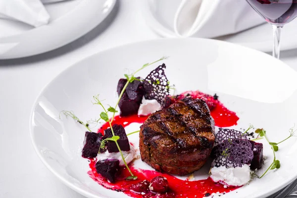 A piece of grilled beef with sprouts, beets, lacy black chips and cherry sauce on a white round plate with a glass of red wine. Horizontal orientation