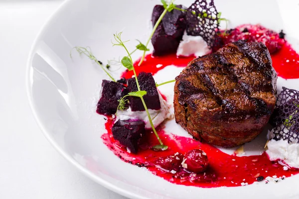 A piece of grilled beef with sprouts, beets, lacy black chips and cherry sauce on a white round plate with a glass of red wine. Horizontal orientation