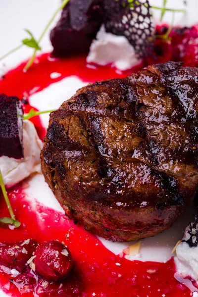 A piece of grilled beef with sprouts, beets, lacy black chips and cherry sauce on a white round plate with a glass of red wine. Vertical orientation