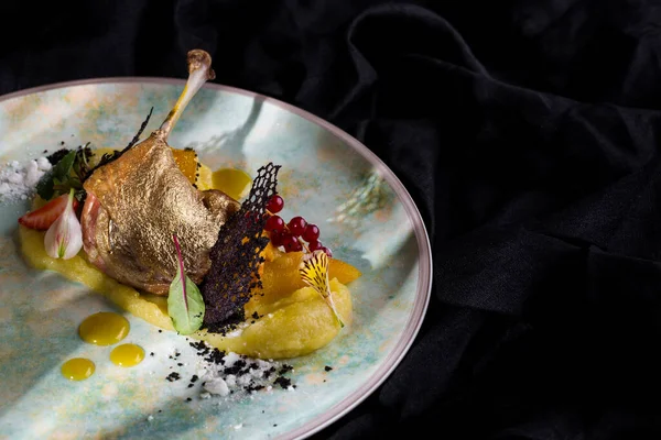 Grilled duck leg with bone, golden skin, red currants, flower petals, powdered sugar, strawberries, mashed potatoes, lace chips, honey mustard sauce on a round, white plate and dark background Horizontal orientation