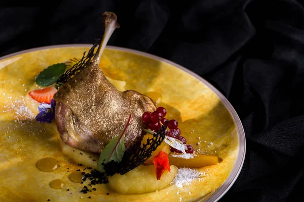 Grilled duck leg with bone, golden skin, red currants, flower petals, powdered sugar, strawberries, mashed potatoes, lace chips, honey mustard sauce on a round yellow plate and dark background Horizontal orientation