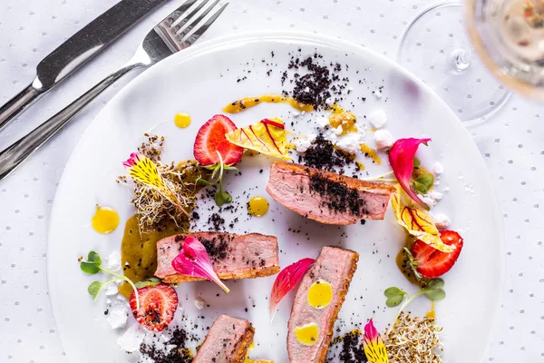 Grilled duck pieces with flower petals, strawberries, sauce, pea sprouts, young shoots, black seasoning on a white round plate with cutlery and a glass of white wine. Horizontal orientation