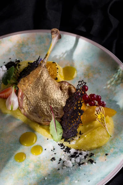 Grilled duck leg with bone, gold rind, red currants, flower petals, powdered sugar, strawberries, mashed potatoes, lace chips, honey mustard sauce on a round, white plate and dark background Vertical orientation