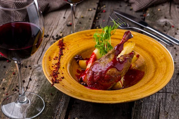 Grilled duck leg on the bone, with jam, green pea sprouts, star anise, pear pieces, black and red peppercorns on a yellow tarkel, with a glass of red wine, with cutlery on a wooden table. Horizontal orientation