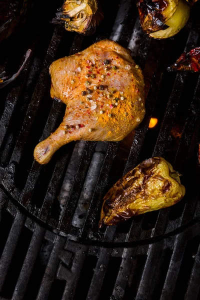 Duck leg with peppercorns and salt, garlic, red pepper and tomato lying on the grill grate over burning coals. Vertical orientation