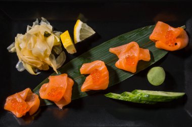 Salmon pieces with red caviar on a leaf with cucumber, wasabi, lemon and ginger on a black background