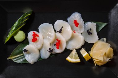Squid pieces with tobiko caviar on a leaf with cucumber, wasabi, lemon and ginger on a black background