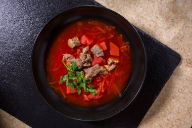 A plate of borscht with beets, meat, parsley, potatoes on a black and white stone background