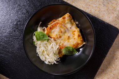Plate of lasagna, eggs, meat, cabbage and basil on black and white stone background