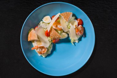 Shrimps with egg, cheese, tomato, bun and lettuce on a blue plate on a black stone background