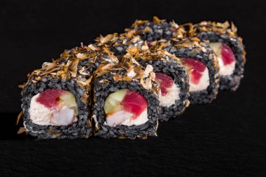 Roll with sprinkles, black rice, nori, cucumber, squid, tuna and cream cheese on a black stone background