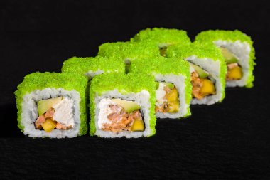 Roll with green tobiko caviar, rice, avocado, cream cheese, fruit and seafood on a black stone background
