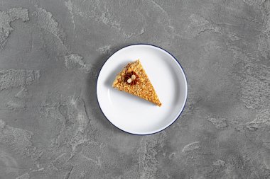 Piece of waffle cake with condensed milk and crushed walnuts. On top is condensed milk and two pieces of peanuts. The cake stands on a white, round, ceramic plate. The plate stands on a gray stone background.