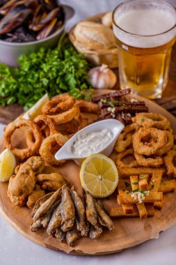 Onion rings in batter, fish in batter, rye croutons with garlic, croutons with white bread, lemon, fried wings, parsley on a board with sauce, a basket of bread, a plate of mussels and a glass of beer. Vertical orientation