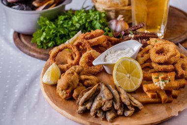 Onion rings in batter, fish in batter, rye croutons with garlic, croutons with white bread, lemon, fried wings, parsley on a board with sauce, a basket of bread, a plate of mussels and a glass of beer. Horizontal orientation