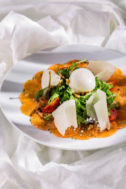 Karışık salatalı somon carpaccio, vişneli domates, ceviz, parmesan dilimleri, kapari ve limonlu şerbet. Yemek yuvarlak, beyaz bir tabakta, tabak hafif, keten kumaş..