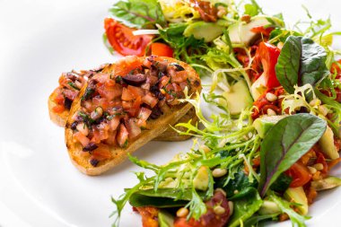 Domatesli, soğanlı, kişniş ve paprikalı bruschetta yanında da domates, çam fıstığı, roka, buzdağı, avokado ve balzamik sos. Yemek beyaz, yuvarlak bir tabakta..